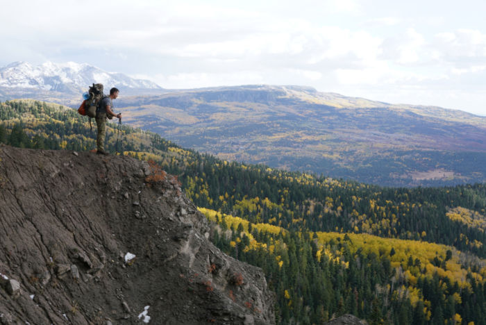 A hiker or hunter stands at the edge of a steep slope with vast views of beautiful mountains.