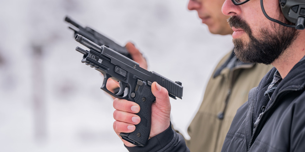 Pistol shooter wearing ear and eye protection, combat gloves and vest, aiming a flashlight mounted pistol with SIG suppressor installed. 