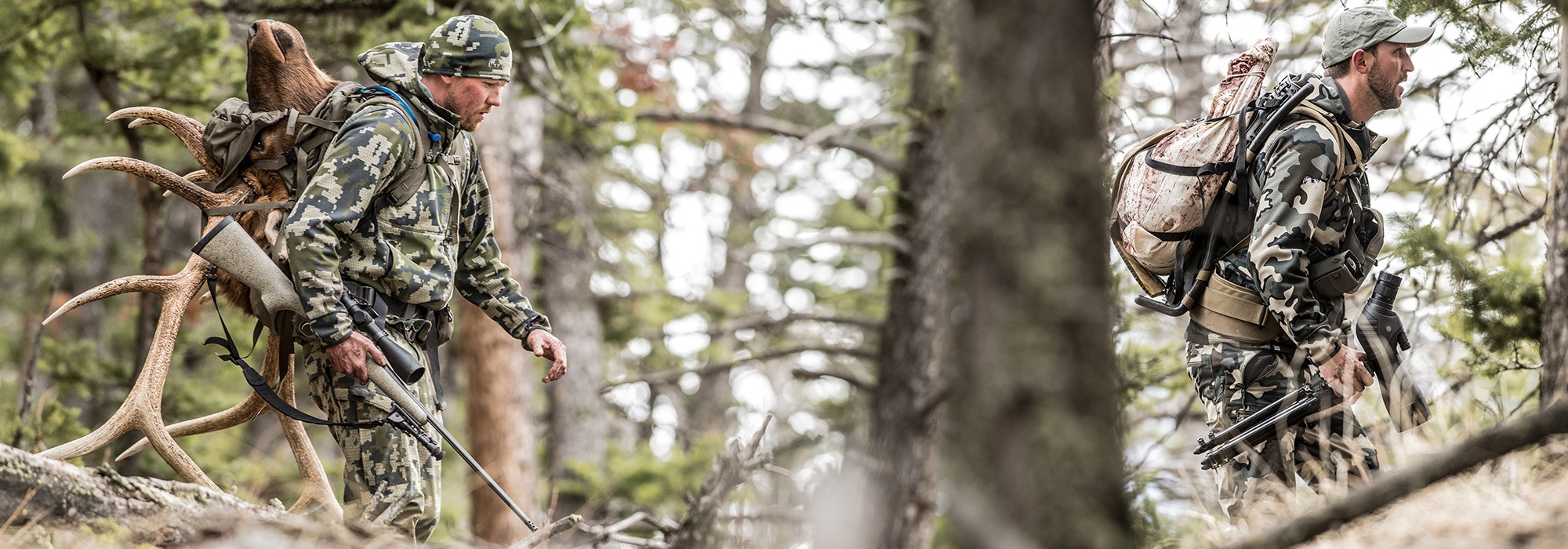 Two hunters in hunting gear with rifle scope, electro optics and binoculars for long range hunting.